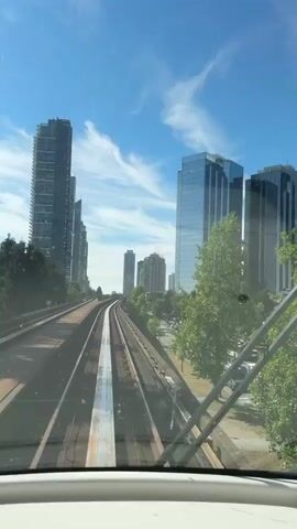 The video shows a view from the front of a train or light rail vehicle, traveling along an elevated track. The train is moving through an urban environment with a clear blue sky and white, wispy clouds. On the left side of the track are several tall, modern skyscrapers, some with glass facades. On the right side of the track, there is a road with cars and trucks visible, as well as trees and some lower-rise buildings. The tracks themselves appear to be well-maintained, with visible metal rails and concrete ties. The view is symmetrical, with the tracks converging in the distance, creating a sense of forward motion. The windshield of the vehicle is visible in the foreground, with two wiper blades crossing the lower part of the frame. The overall impression is one of efficient urban transit moving through a modern city on a bright, sunny day.