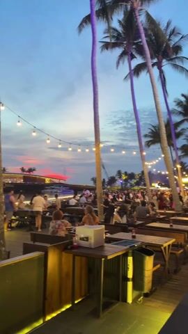 The video shows an outdoor restaurant at dusk with many patrons seated at wooden tables and benches. The restaurant is decorated with string lights and surrounded by palm trees, some of which are lit with purple lights. The restaurant deck is elevated and overlooks a body of water. A bridge with blue lights can be seen in the distance. Some people are standing and walking through the restaurant, while others are seated and eating or talking. A waiter in a yellow and red shirt is visible. A couple in formal attire is seated at a table with drinks. A trash can and a box are near the edge of the deck. The sky is a mix of blue, orange, and pink, suggesting the sun is setting.