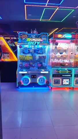 A man wearing a black t-shirt with a logo on the back and a baseball cap walks through an arcade. The arcade is dimly lit and filled with rows of brightly lit claw machines, each containing plush toys. The floor is tiled, and the ceiling is adorned with colorful neon lights in a grid pattern. The claw machines have illuminated buttons and displays. The man is walking away from the camera and towards the right side of the frame.