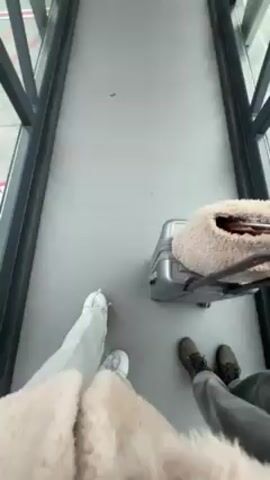 The video shows a first-person view of someone walking along an airport jet bridge with a suitcase, followed by a quick selfie of the person and a man walking together. The walkway has grey flooring, metal framed windows on both sides, and advertisements on the walls. The person filming is wearing a light-colored, fuzzy coat, light jeans, and white sneakers, while the man beside them wears glasses, a grey scarf, and a dark coat.