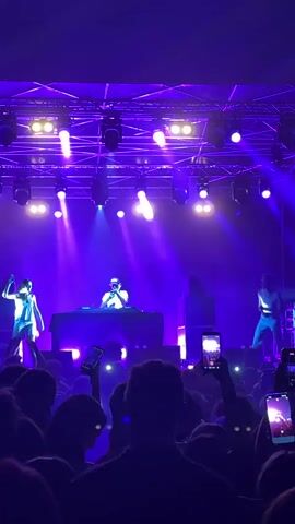 The video shows a concert with a stage illuminated by purple lights. On stage, there are at least three people. One person is standing behind a table, appearing to be a DJ, wearing a white shirt and a hat. Another person, dressed in a light-colored outfit, is standing to the left of the DJ. A third person, in a white shirt and dark pants, is standing to the right of the DJ, facing the audience. The audience is visible in the foreground, with many people holding up their phones to record. The atmosphere appears to be energetic and engaged.