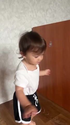 A toddler with brown hair and blue eyes, wearing a white bodysuit and black shorts with white stripes, is dancing. The toddler is in a room with a textured wallpaper and a wooden floor. The toddler is moving their arms and head rhythmically. The video captures the toddler's playful and energetic movements.