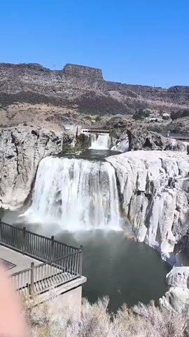 The video shows a wide waterfall cascading down a rocky cliff under a clear blue sky. A paved viewing area with a wooden railing is in the foreground to the left. The water is a deep green, and mist rises from the base of the falls. In the distance, there is a smaller waterfall and a bridge crossing the river above the main falls. The surrounding landscape is arid and rocky, with sparse vegetation.