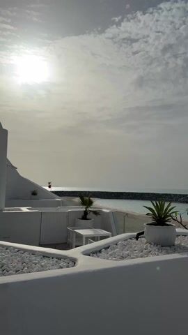 From an elevated perspective, the camera captures a daytime beach scene under a sky obscured by thin, mottled clouds and a bright sun. The foreground is dominated by white-painted architectural structures, including a raised parapet adorned with rounded white stones. Three potted plants add greenery, with two smaller palm trees and one succulent-like plant. A simple white table and a section of white fencing are also visible.

Beyond the white structures, a sandy beach stretches to meet the ocean. A dark rock barrier extends into the water, creating a protected lagoon. In the distance, a red flag is visible. The water is calm, reflecting the sunlight with a shimmering glare.