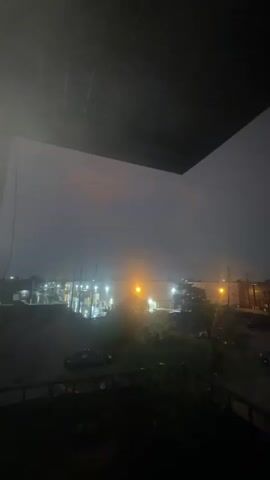 The video captures a stormy sky over a semi-urban landscape. The sky is dark and cloudy, with visible rain falling. Streetlights and building lights cast an orange glow through the rain. Cars are parked on the side of the road, and some are on a wet-looking street. The foreground shows a dark railing and a dark area that could be a balcony or porch.