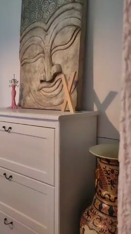 The video pans across a room, focusing on a white dresser with three drawers. On top of the dresser is a large, carved relief of a Buddha's face, a small pink jewelry stand, and a wooden letter "X". To the right of the dresser, a large ornate vase with intricate patterns is visible. To the left of the dresser, a window is covered by sheer white curtains and dark green velvet drapes. A wooden table with a dark metal base is partially visible in the foreground, near the window. The room appears to be a bedroom or living area with a calm, decorative aesthetic.