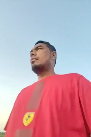 A medium-skinned man with a beard and a red t-shirt is looking up at the sky. He is facing the camera and then looks to his right and back to the camera. The sky is a pale blue, bordering on white. The man's shirt has a dark brown stripe down the center and a yellow and black logo on the bottom left.