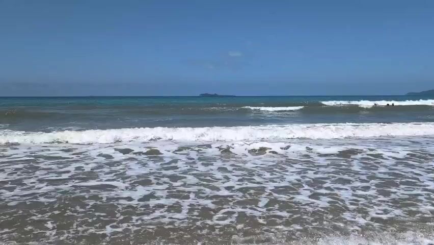 The video shows a serene ocean scene with clear blue skies. Gentle waves roll towards a sandy beach, creating foamy white crests as they break. In the distance, several small islands are visible on the horizon, adding depth to the landscape. A few people can be seen swimming and enjoying the water, their figures silhouetted against the bright sea. The overall atmosphere is one of tranquility and relaxation, characteristic of a beautiful beach day.