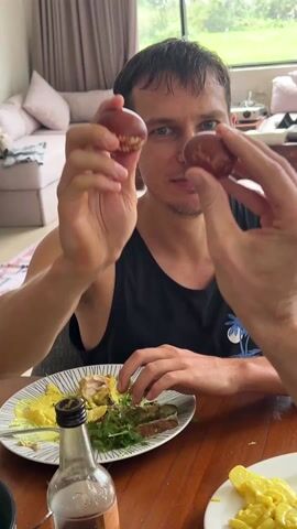 The video shows a fair-skinned man with short brown hair and blue eyes sitting at a wooden table and holding up two small, reddish-brown objects in front of his face. He is wearing a black tank top with a blue palm tree design on it. A plate of food is visible on the table in front of him, containing scrambled eggs, greens, and what appears to be meat. A small bottle with a dark liquid is also on the table. The man is smiling and appears to be engaged with the person filming. The background includes a living room setting with a light-colored sofa, pillows, and a window with curtains. The scene appears to be indoors, possibly in a home or dining area. The focus is on the man and the objects he is holding, with the camera positioned relatively close.