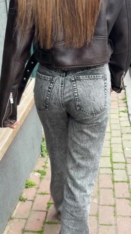 A rear-view shot follows a person walking on a brick-paved walkway. The person has long brown hair and is wearing a brown leather jacket and gray jeans with white sneakers. The person is walking away from the camera. Two people walking in the same direction pass by. The walkway has green vegetation growing in the cracks and a green door and a wooden bench can be seen to the left.