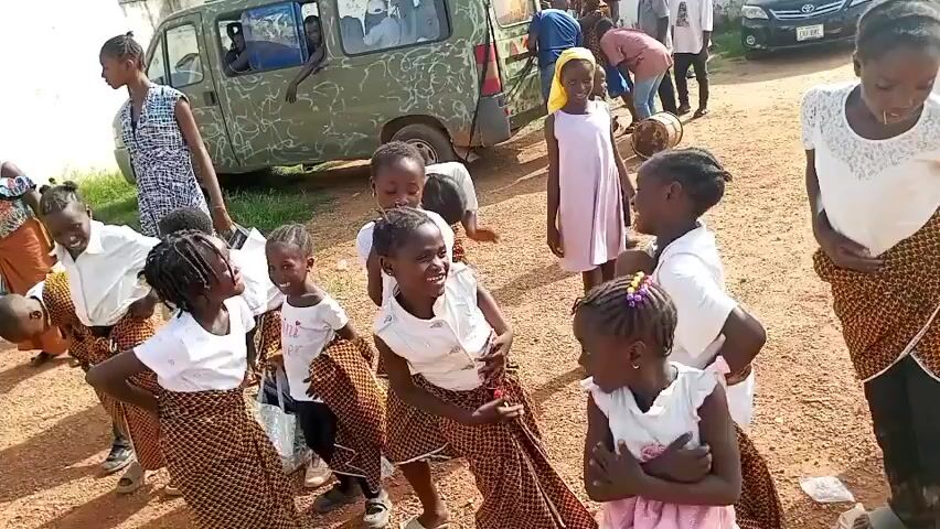 A group of young children, mostly girls, are gathered outdoors. They appear to be dressed in similar outfits consisting of white tops and patterned wraps or skirts. The children are interacting with each other, smiling, posing, and possibly dancing. In the background, there are adults, a vehicle, and other children. The ground appears to be dirt or reddish soil. The overall atmosphere seems cheerful and celebratory.