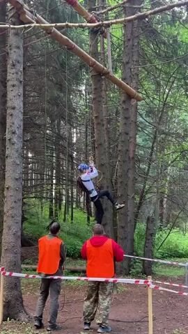 A young person wearing a helmet and a harness is suspended in the air on a rope, climbing an obstacle course in a forest. The course consists of wooden beams and ropes strung between tall trees. Two adults, wearing orange safety vests, stand below, watching the climber. The ground is covered in dirt and pine needles. The trees are tall and densely packed, creating a shaded environment. The overall scene depicts an outdoor recreational activity, likely a ropes course or adventure park.