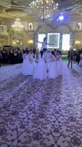 A group of young women in white dresses are dancing in a circle in a large, ornate ballroom. The woman in the center is standing on a raised platform and is also wearing white. The other women are holding long, white ribbons and are moving them in unison. The ballroom has a large chandelier, and there are many guests watching the performance.