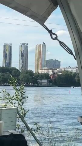 The video shows a view across a body of water towards a city skyline. In the foreground, the edge of a white canopy is visible, with metal cables and turnbuckles attached. To the left, a white paper cup and a dark object, possibly clothing, are on a table. A small green plant is also in the foreground. The water is rippling. In the middle ground, there is a line of trees and some low buildings. In the background, several tall, modern apartment buildings rise into the sky, some with colorful facades. A person on a jet ski is visible on the water, leaving a wake and moving to the right side of the frame. The sky is partly cloudy.