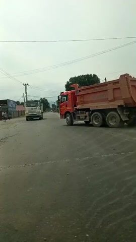 The video begins by showing a street scene with multiple vehicles. A large orange dump truck is on the right side of the road, facing away from the camera. A white truck is further down the road. A black car is parked on the left. Traffic lights are visible, with the red light illuminated. The road surface appears worn and uneven.

The scene then shifts to show a street with buildings. On the left, there is a building with a blue and silver facade and a sign that reads "КЕТМАН.KG". Next to it is a building with a red roof and signage that includes "НЕМАН АПТЕЧНЫЙ ПУНКТ". Three people are walking on the sidewalk in front of this building, two adults and a child.

The video continues, showing the "НЕМАН" building more clearly. Two people are walking away from the camera on the sidewalk.

Another building is visible, with a yellow awning over an entrance. A silver car is parked in front of it. To the right of this building is a blue sign with white text that appears to read "СЕМЕЙКА".

Finally, the video shows a two-story building with a blue, red, and white patterned roof. There is a sign on the lower level that reads "ШАУРМА КРЫЛЫШКИ САМСА". Two outdoor grills are visible to the left of this establishment. The overall environment appears to be a commercial area with shops and restaurants. The sky is overcast.