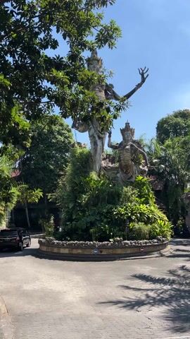 The video presents a daytime view of a large stone sculpture, possibly in Bali or another Southeast Asian country, situated on a traffic circle. The sculpture depicts two figures, one taller than the other, adorned with what appear to be traditional headdresses or crowns. They are surrounded by lush green vegetation and foliage.  The base of the sculpture sits on a circular structure, which is inscribed with text that reads "The Royal Pita Maha Ubud". A black SUV is visible moving around the traffic circle to the left of the sculpture, and other vehicles are parked in the background. The sky is clear and blue, with some trees providing shade to the left of the frame. The paving of the traffic circle is made of interlocking concrete blocks.