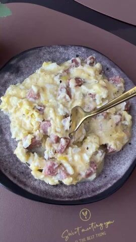 A close-up shot shows a plate of scrambled eggs with sausage. A gold fork lifts a portion of the food, revealing strands of melted cheese. The plate is gray with a black rim and sits on a brown placemat with the words "Sweet meeting you" printed on it. A portion of a dark brown or black surface is seen at the top of the frame. A small potted plant is visible in the background.