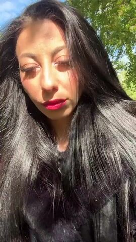 A woman with long, dark hair and red lipstick faces the camera. She is wearing a black top or jacket. Behind her, there is green foliage and a blue sky. The sunlight shines on her hair and face.