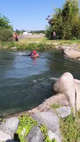 A person is navigating a man-made rapid on a river. They are on a red inflatable raft, wearing a helmet.  The river has concrete structures creating the rapids. The riverbanks are lined with trees and vegetation. On the far bank, there is a building and a few parked vehicles visible. The sky is blue, indicating a sunny day.  Another person on a raft is visible in the background. The camera is stationary, providing a steady view of the scene.