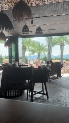 The media file depicts a view from an indoor seating area looking out onto a resort or pool area. In the foreground, dark, intricately carved chairs are visible, suggesting a stylish lounge or restaurant setting. Hanging above, decorative lamps with woven shades add to the ambiance.

Through a large glass opening, the scene transitions to an outdoor, sunny environment. Several people are lounging by a pool, some on sunbeds and others in beanbag chairs. Palm trees are a prominent feature, with their tall, textured trunks and lush green fronds reaching towards a bright blue sky with scattered clouds. In the distance, rolling hills or mountains form a scenic backdrop, dotted with trees and possibly some buildings. The weather appears to be warm and sunny, indicative of a pleasant day.

The people in the pool area are casually dressed, some in swimwear, and appear to be relaxed and enjoying the leisure atmosphere. A young boy in swim trunks is visible, holding what looks like a toy. The overall impression is that of a relaxed vacation or resort setting on a clear, bright day. There is no visible text within the media.