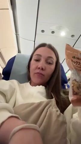 A woman sits in an airplane seat and holds a bag of mixed nuts in front of her face. She looks directly at the camera and shakes the bag. She is wearing a white sweater and has long brown hair. The seat behind her is blue. The airplane interior is visible in the background.