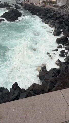 The video shows a scenic view of the ocean in Puerto de la Cruz, Spain. The choppy, turquoise water crashes against dark volcanic rocks that line the coastline. Waves create white foam as they surge and recede. In the distance, a promenade with people can be seen along the top of the rocky shore. The weather appears to be overcast or hazy, suggesting it might be morning or a cloudy day. The overall mood is one of natural beauty and the power of the sea. There are no discernible objects or text within the frame.
