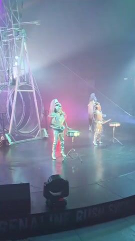 The video displays a stage performance with theatrical lighting. In the foreground, two individuals are positioned at separate drum sets, dressed in silver costumes and elaborate headpieces. To the left, a large metallic structure, possibly part of a Ferris wheel or a similar apparatus, is visible. The stage floor is dark and reflective, with several stage lights positioned on the ground. The background features an audience area, with rows of seats and people visible, though their faces are indistinct due to the distance and lighting. The overall atmosphere is energetic and visually striking, with colored lights and haze contributing to the theatrical effect. A banner along the bottom edge of the frame reads "ADRENALINE RUSH SHOW".