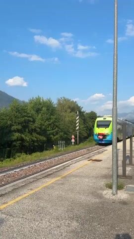 A train with a green and blue exterior approaches a station. The train is traveling on the right side of the frame. The station platform is grey with a yellow line along the edge. The train is on the left side of the platform. Trees and mountains are visible in the background. The sky is blue with a few white clouds. A tall pole is in the foreground on the right side of the frame.