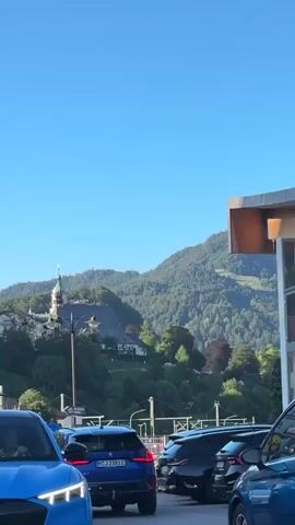 A blue car is parked in the foreground. Behind it are several other cars, including a black SUV. In the background is a city with buildings and a church on a hill. The sky is clear and blue.