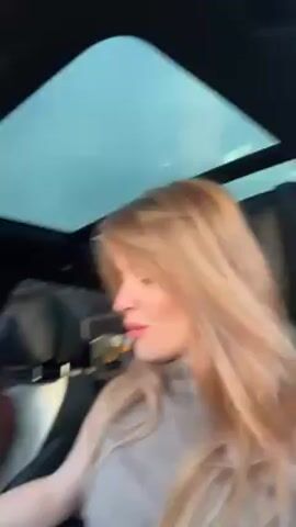 Inside a car, a woman with long blonde hair, wearing a gray sleeveless turtleneck, smiles at the camera and says, “What’s up guys. Let’s take a ride.” The car starts. The car’s interior is black and white. The camera pans to the driver who has curly brown hair and is wearing a brown jacket. The dashboard is visible and has a white steering wheel.  Outside, other cars are parked along a street.