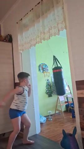 A young boy is practicing martial arts in a hallway. He is wearing a white tank top with a blue print and blue shorts. He is barefoot and stands on a dark wooden floor. In front of him is a doorway, beyond which is a room with light green walls. A punching bag hangs in the center of the doorway, swinging. The boy is in mid-motion, appearing to be throwing a punch or kick at the bag. The punching bag is black with red accents and a graphic of what appears to be a red car. In the background of the room, a child's easel with drawings is visible, as well as a small Christmas tree decorated with colorful ornaments and lights. A white-tasseled curtain hangs across the top of the doorway. The lighting is bright, suggesting daytime.