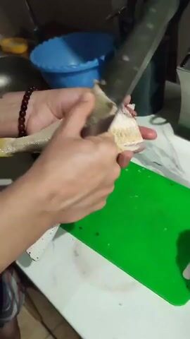 The video shows a person's hands preparing a fish. The person is using a knife to remove the skin from the fish, which is placed on a green cutting board. The fish appears to be raw and is being held by the tail. There are also other kitchen items visible in the background, such as bowls and utensils. The overall scene suggests a domestic kitchen environment where food preparation is taking place.