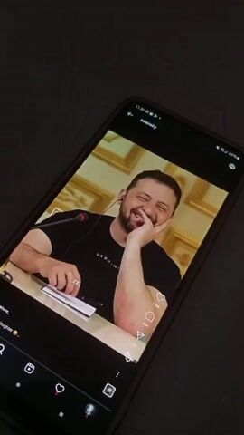 A person holds a phone displaying an Instagram post of Volodymyr Zelenskyy. In the first frame, Zelenskyy is wearing a black shirt with the word 'Ukraine' on it and is smiling and leaning on his hand. Subsequent frames show different videos of Zelenskyy. The phone is lying on what appears to be a patterned surface, possibly a bedspread or sofa. The room is dimly lit. The phone's interface shows typical Instagram features, like the heart icon, comment icon, and share icon.
