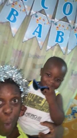 A blurry video features two individuals. One, a dark-skinned woman, is wearing a silver tinsel crown and a yellow shirt. She is smiling and appears to be holding the camera. Next to her is a young, dark-skinned child in a yellow and white tank top, with blue sunglasses resting on the front of the shirt. A banner with blue letters that spell "WELCOME BABY" hangs in the background against a green and yellow patterned curtain. The camera moves around during the video.