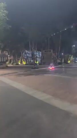 At night, a small, illuminated red vehicle is visible on a concrete plaza lined with palm trees. The vehicle is moving away from the viewer. In the background, there's a structure that looks like a small village or playground. The lighting is dim, with string lights woven through the trees. The camera pans slightly to follow the vehicle as it moves.