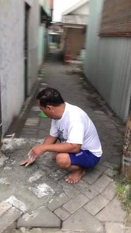 The video shows a man in a white t-shirt and blue shorts squatting on a cobblestone path next to a building. He is facing away from the camera. To his left is a closed metal gate with vertical bars and a decorative top. To his right are two large, dark vertical poles with electrical wires wrapped around them. The ground appears to be a mix of cobblestones and dirt, with some debris scattered around. The man appears to be looking down at something on the ground. The lighting suggests it is daytime, possibly overcast.