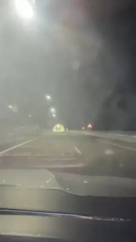 Driving down a highway at night, the view from inside the car shows a tunnel ahead, illuminated with bright yellow and green lights. Streetlights line the road, and the visibility is somewhat obscured, possibly due to rain or fog. A traffic sign is visible on the right side of the road, and the car's dashboard is in view. The road markings are clear.