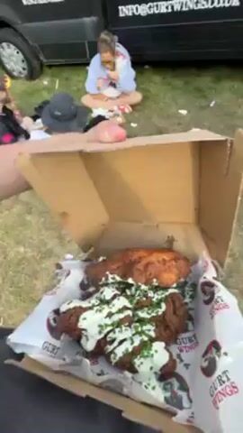 Here is a detailed description of the video:

**Overall Impression:** The video showcases a food vendor, Gurt Wings, at an outdoor event, likely a festival or fair. People are milling around, enjoying food and the atmosphere.

**Setting:** The scene is set in an open, grassy area, possibly a field. The ground is covered with grass and some litter. In the background, there are trees and other tents or structures that suggest a larger event. The sky is blue with scattered clouds.

**People:**
*   Several people are visible, including customers and staff around the food vendor.
*   Some individuals are wearing casual summer clothing, like shorts and t-shirts.
*   One person is shown holding a box of chicken wings, which seems to be the focus of the video.
*   People are seen standing, walking, and sitting on the ground.

**Objects:**
*   A black food truck or van with "Gurt Wings" branding. The side of the truck displays the company's website and contact information.
*   The food truck has an open serving area where staff can interact with customers.
*   Boxes of food, specifically chicken wings, are featured. The wings appear crispy and are topped with a sauce and garnish.
*   A flag, or banner, hangs near the food truck.
*   Other items like hats, bags, and discarded food containers are scattered in the background.

**Actions:**
*   The camera person holds and displays a box of chicken wings.
*   Customers approach the food truck, suggesting they are ordering or receiving food.
*   People are interacting and chatting with each other.
*   Individuals are moving around within the area, likely exploring the event.

**Interactions:**
*   There is an interaction between customers and the food truck staff.
*   People are talking and interacting with each other, possibly related to the food or the event.

In summary, the video shows a lively scene around a food vendor at an outdoor event, emphasizing the food available and the overall festive atmosphere.