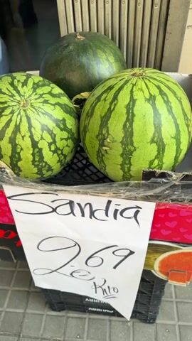 The media file captures a street-side display of watermelons, likely outside a grocery store or market stall in L'Hospitalet de Llobregat, Spain. The scene is brightly lit, indicating daytime conditions.

In the foreground, several large, round watermelons are arranged in a black plastic crate, which is placed inside a decorative cardboard box. The cardboard box features a red rim adorned with white hearts and floral patterns. Two watermelons are most prominent, showcasing distinct green rinds with darker stripes and mottled patterns. A third, slightly darker green watermelon is visible behind them, positioned further back in the display.

A handwritten white paper sign is taped to the front of the cardboard box. The sign clearly displays "Sandia" (Spanish for watermelon) at the top in cursive script. Below this, the price "2.69" is written, followed by "Kilo," indicating a price of 2.69 per kilogram. Partially visible, upside-down text on the black plastic crate includes "CHURRY" and "ABBIU" (possibly part of "ABBIUS").

The background features a partially open roll-down metal security gate or shutter, suggesting a storefront entrance. The ground is paved with square, light-colored tiles, characteristic of a pedestrian street or sidewalk. No people are visible in the frame, but the arrangement and pricing of the fruit indicate a commercial transaction setting.