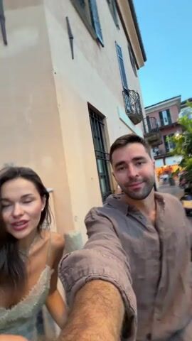 The video begins with a close-up selfie of a young couple. The woman has dark hair and a light-colored top, and she is making a surprised facial expression. The man has dark hair and is wearing a tan button-down shirt. He is smiling at the camera. In the background, there is a building with windows and a lamppost. Another person can be seen in the background. The scene appears to be outdoors, possibly at a restaurant or cafe, with people seated at tables. The camera then pans slightly, revealing more of the surroundings, including an Italian flag on a building. The scene is bustling with activity, with many people visible. The video then abruptly blurs and shifts focus, showing a more abstract view of the environment before returning to a shot of the couple. The woman’s surprised expression is more pronounced, and she is now looking directly at the camera. The man is still smiling. The Italian flag is still visible in the background, along with a crowd of people. The video ends with a shot of the couple from behind, sitting on a wall and looking towards the crowd. The lighting suggests it is late afternoon or early evening.