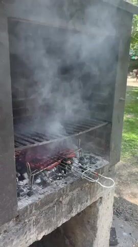 The video shows a close-up of a grill with meat cooking over charcoal. Smoke billows from the grill. In the foreground, two people are preparing raw meat. A red bag with the word "CARBON" and "CARVAO" printed on it is visible behind the meat. The environment appears to be outdoors, with trees and other people visible in the background.