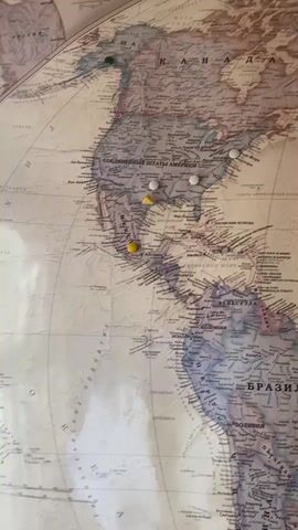 The video displays a world map on a wall, likely a map used for travel tracking. The map is covered in various colored pushpins, each marking a different location. The pins are concentrated in North America, Europe, Asia, and Africa, suggesting extensive travel. The map is framed and appears to be made of a reflective material, possibly plastic laminate, which causes some glare and obscures certain details. The camera pans across the map, offering a view of the pins and the geographical locations they represent. The primary subject is the world map and the pushpins, with no people or other significant objects visible in the frame. The environment is implied to be indoors, given the presence of the framed map on a wall. The purpose of the pushpins is to indicate places visited by the subject's husband, as stated in the Russian audio.
