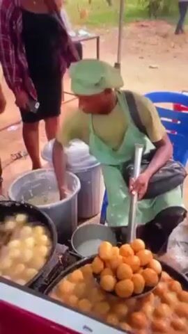 A person wearing a green apron and hat is shown working at an outdoor food stall. They are seated on a blue chair and are using their hands to scoop batter from a grey bucket into a pot of hot oil where round, donut-like pastries are frying. Another pot next to it is filled with already cooked, golden-brown pastries. There are other people standing around watching, and in the background, a table with more people can be seen. The stall appears to be set up on a dirt surface.
