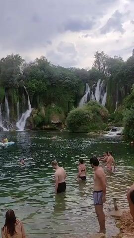 The video shows a scenic view of waterfalls cascading into a clear, green body of water. Several people are swimming and relaxing in the water. In the background, there are lush green trees and a cloudy sky. Some people are gathered on the shore, enjoying the view and the atmosphere.