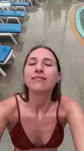 A woman in a brown bikini stands outside near a pool, facing up and smiling as a wave of water drenches her. The background includes a gray patio, a pool, and some blue lounge chairs. The weather appears to be wet.