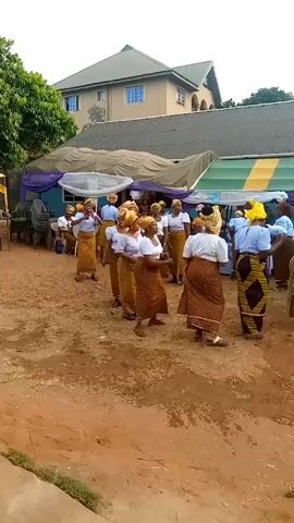 A group of about twenty women, all dressed in similar clothing, dance in an open dirt space. The women are all wearing white blouses and skirts with brown patterns, and most of them are also wearing yellow head coverings. They are moving as a group and appear to be having fun. In the background, there is a large house with a green roof and a covered area with chairs and decorations.