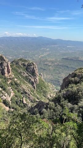 The video shows a panoramic view of the Montserrat mountain range in Spain, under a blue sky with scattered clouds. The foreground features rugged, rocky terrain covered in green trees and shrubs. In the distance, a valley stretches out, dotted with small towns and patches of agricultural land. The camera slowly pans to the left, revealing buildings built into the side of the mountain, including what appears to be a monastery complex. A path runs along the mountainside, connecting the different structures. The overall impression is one of natural beauty combined with human construction, showcasing the unique landscape of Montserrat.