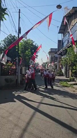 A group of young people, likely students, in uniform are marching down a street decorated with Indonesian flags. The flags are red and white, the colors of the Indonesian flag, and are strung across the street and along the sides. The students are wearing matching outfits, some with red tops and black pants, and others with white tops and black pants, with some wearing headscarves. They appear to be participating in a parade or a patriotic event. The street is paved and lined with houses and trees. The sky is clear and blue, indicating a sunny day. The perspective is from the ground, looking up the street.