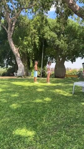 Two children are playing in a grassy area with large trees in the background. A boy in blue shorts stands facing right. A girl in a swimsuit is facing away from the camera and appears to be bending over or getting ready to move. The girl then walks to the right and turns to face the boy. There are chairs and a trash can visible in the background.