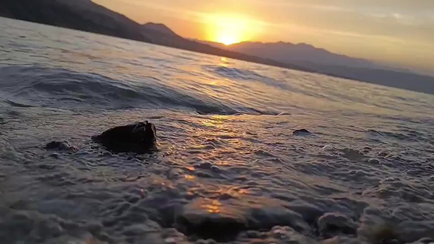 A low-angle shot captures gentle waves lapping at a rocky shore during sunset. The sun, a bright orb, is low on the horizon, casting a warm glow across the water and silhouette mountains in the distance. Small pebbles and rocks are visible at the water's edge, with some foam from the waves accumulating around them. The water is choppy, creating small ripples and waves that break softly on the shore. The overall scene is peaceful and serene, with the colors of the sunset dominating the view.